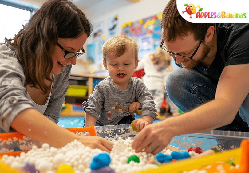 A toddler smiling while playing with a colorful sensory bin filled with rice, scoops, and toys, engaging in hands-on learning through touch and exploration.