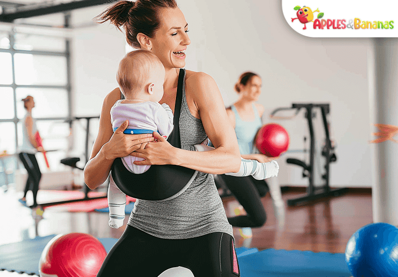 A smiling mother doing squats while holding her baby, engaging in a fun bonding workout session at home.